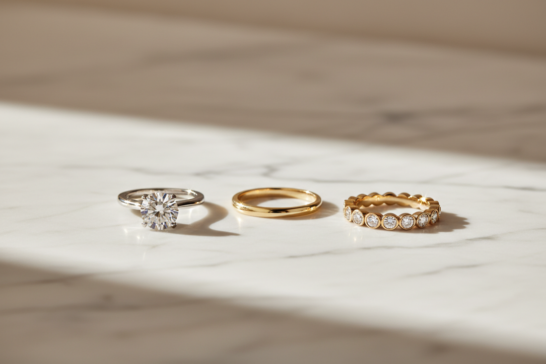 Three fine jewelry rings on white marble — platinum solitaire engagement ring, 18K gold wedding band, and diamond eternity anniversary band — representing the three milestones of love, by JewelryRich Fine Jewelry