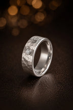 18K white gold hammered brushed finish men's wedding band with single bezel-set diamond accent, flat lay on dark surface